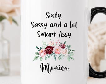 May include: A white ceramic mug with a black handle and rim. The front features the black text "Sixty, Sassy and a bit Smart Assy" above a floral illustration of pink and dark red roses with green leaves, followed by the name "Monica" in black.