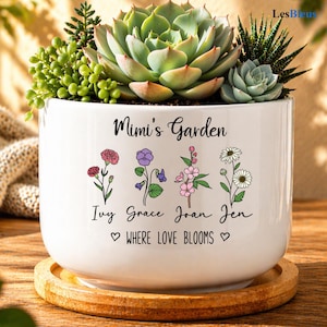 May include: White ceramic planter with a variety of succulents and the text "Mimi's Garden". The planter features floral illustrations and the names Ivy, Grace, Joan, and Jen, with the phrase "WHERE LOVE BLOOMS". The planter sits on a wooden coaster.