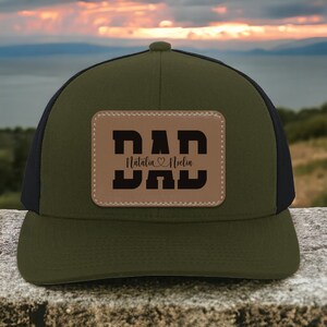 K&ouml;nnte beinhalten: Eine gr&uuml;ne und schwarze Trucker-Kappe mit einem braunen Leder-Patch, auf dem "DAD" steht, darunter die Namen "Natalia" und "Noelin".