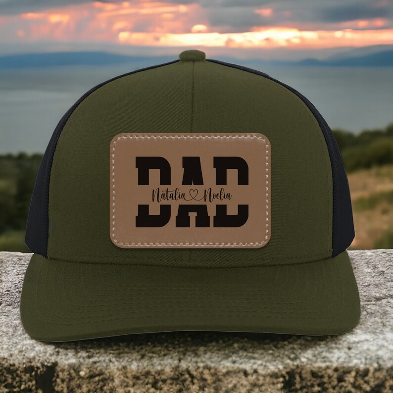 K&ouml;nnte beinhalten: Eine gr&uuml;ne und schwarze Trucker-Kappe mit einem braunen Leder-Patch, auf dem "DAD" steht, darunter die Namen "Natalia" und "Noelin".