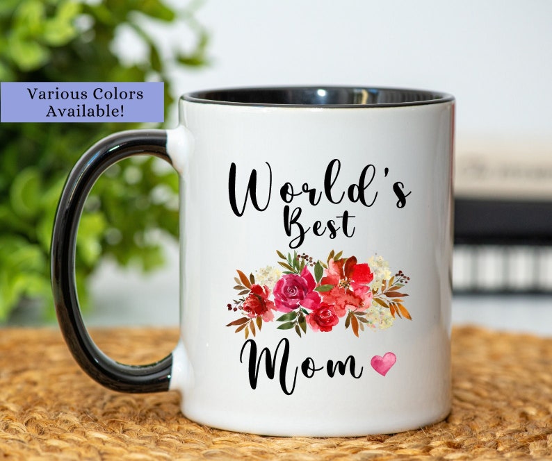 May include: White ceramic mug with a black rim and handle. The mug has a floral design and the text "World's Best Mom" with a small pink heart.