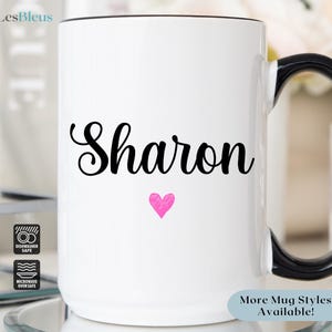 Puede incluir: Taza de cer&aacute;mica blanca con asa negra. La taza tiene el nombre "Sharon" escrito en letras cursivas negras con un coraz&oacute;n rosa debajo del nombre. La taza es apta para lavavajillas y microondas.