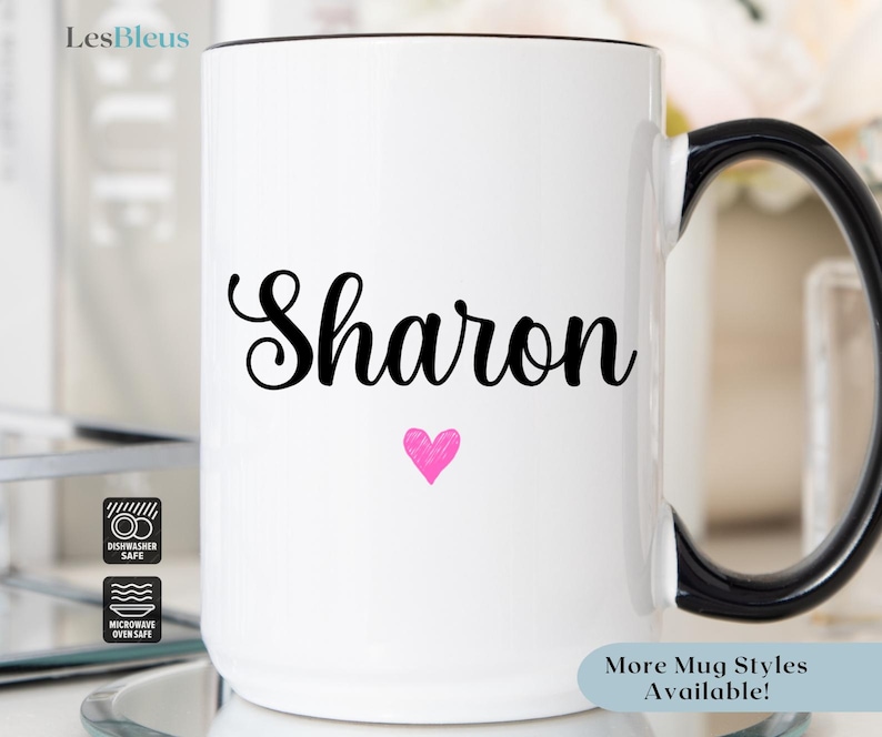 Puede incluir: Taza de cer&aacute;mica blanca con asa negra. La taza tiene el nombre "Sharon" escrito en letras cursivas negras con un coraz&oacute;n rosa debajo del nombre. La taza es apta para lavavajillas y microondas.