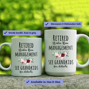 May include: Two white ceramic mugs with black text that reads "Retired Under New Management See Grandkids for details". The mugs have a floral design with pink, red, and white flowers. The text is on a white background. The mugs are microwave and dishwasher safe.