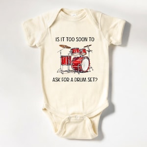 May include: A cream-colored baby onesie with a black and white graphic of a drum set and the text "Is it too soon to ask for a drum set?"