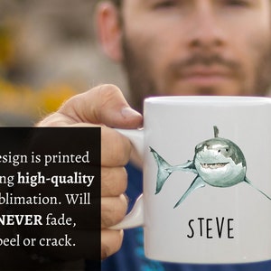 May include: White ceramic mug with a watercolor illustration of a great white shark and the name "STEVE" printed in black below the shark.