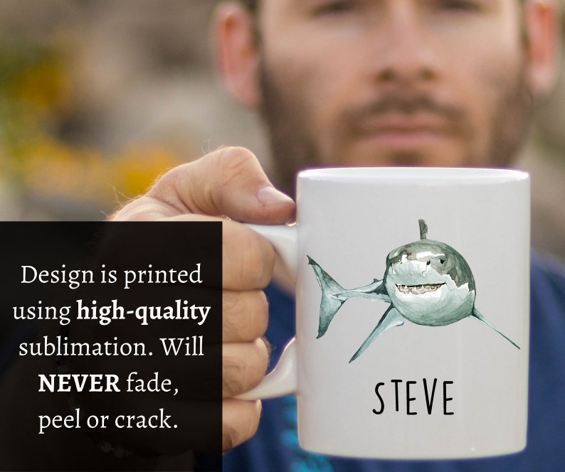 May include: White ceramic mug with a watercolor illustration of a great white shark and the name "STEVE" printed in black below the shark.