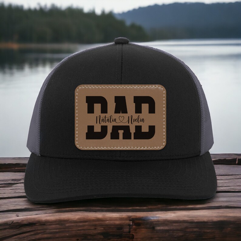 K&ouml;nnte beinhalten: Schwarze und graue Trucker-Kappe mit einem braunen Leder-Patch, auf dem "DAD" steht, darunter die Namen "Natalia" und "Noelle".