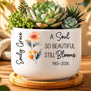 May include: White ceramic planter with a variety of succulents and greenery. The planter features the text "Emily Grace" and "A Soul So Beautiful Still Blooms. 1965-2026" with floral accents. The planter sits on a wooden coaster.