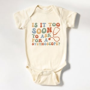May include: A cream-colored baby onesie with the text "Is it too soon to ask for a stethoscope?" in colorful retro-style letters. A red stethoscope is pictured next to the text.