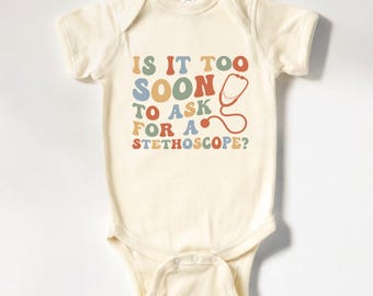 May include: A cream-colored baby onesie with the text "Is it too soon to ask for a stethoscope?" in colorful retro-style letters. A red stethoscope is pictured next to the text.
