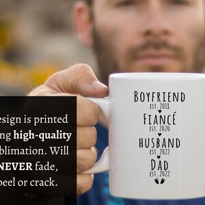 May include: White ceramic mug with black text that reads "Boyfriend Est. 2018, Fianc&eacute; Est. 2020, Husband Est. 2022, Dad Est. 2022" with a heart symbol between each line and a footprint symbol below the last line.
