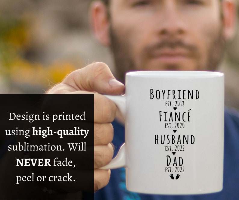 May include: White ceramic mug with black text that reads "Boyfriend Est. 2018, Fianc&eacute; Est. 2020, Husband Est. 2022, Dad Est. 2022" with a heart symbol between each line and a footprint symbol below the last line.
