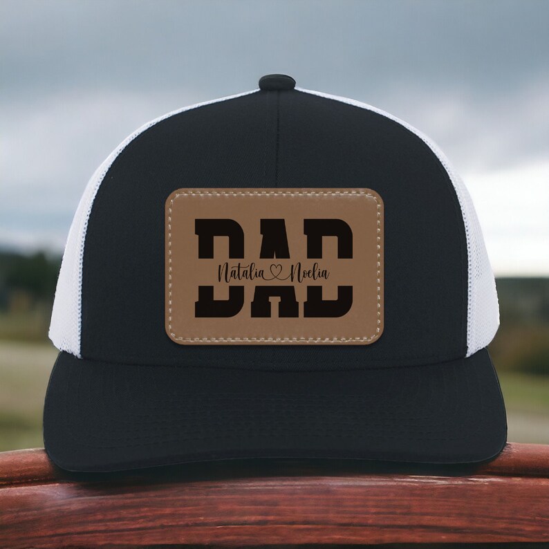 K&ouml;nnte beinhalten: Schwarz-wei&szlig;e Trucker-Kappe mit einem braunen Leder-Patch, auf dem "DAD" steht, darunter die Namen "Natalia" und "Noela".