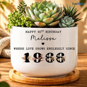 May include: A white ceramic planter with a wooden base, filled with various succulents. The planter has the text "HAPPY 60TH BIRTHDAY Melissa" and "WHERE LOVE GROWS ENDLESSLY SINCE 1966".