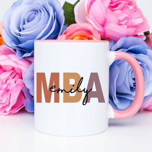 Może przedstawiać: Biały kubek ceramiczny z r&oacute;żowym rantem i uchwytem. Kubek ma nadrukowany tekst "MBA emily" brązowymi i r&oacute;żowymi literami.