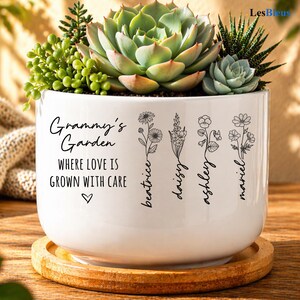 May include: A white ceramic planter with the text "Grammy's Garden" and floral illustrations. The planter holds various succulents and sits on a wooden coaster. The text "WHERE LOVE IS GROWN WITH CARE" is also included.