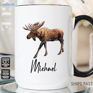 May include: White ceramic mug with a black handle. The mug features a watercolor illustration of a brown moose with large antlers. The name "Michael" is printed in black cursive below the moose. The mug is dishwasher and microwave safe.