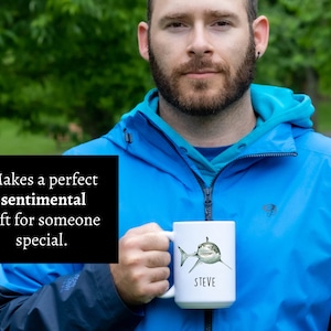 May include: White ceramic mug with a graphic of a great white shark and the name "STEVE" printed on the front. The mug is being held by a person wearing a blue jacket.
