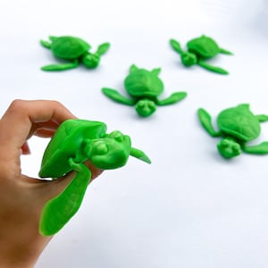 May include: Five green plastic sea turtle figurines on a white background. One turtle is being held in a hand.