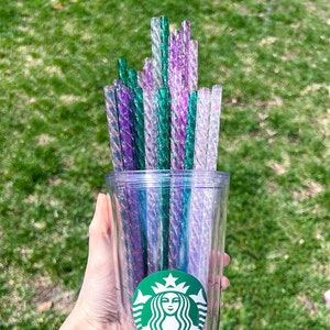 May include: A clear reusable tumbler with a green Starbucks logo and a mermaid design. The tumbler is filled with a bundle of iridescent green and purple straws.