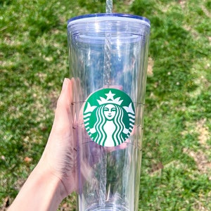May include: A clear plastic tumbler with a green Starbucks logo and a clear plastic straw. The tumbler has a lid and a measurement scale on the side.