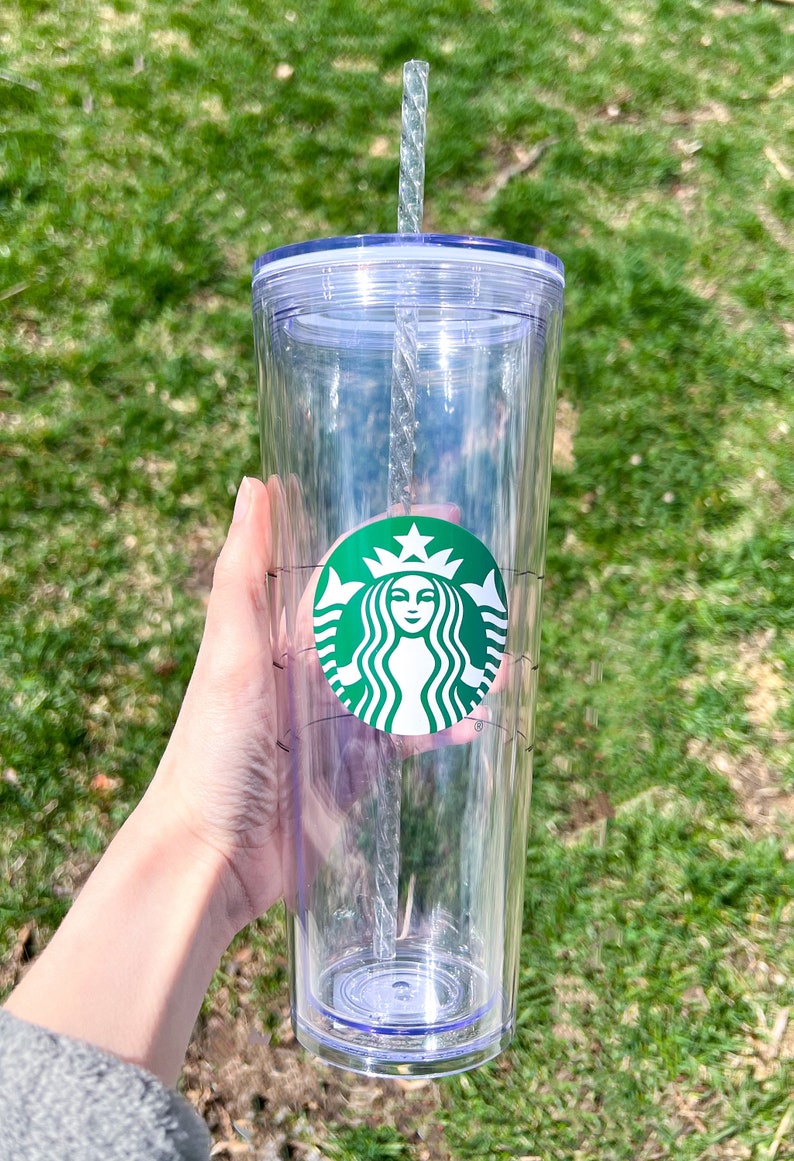 May include: A clear plastic tumbler with a green Starbucks logo and a clear plastic straw. The tumbler has a lid and a measurement scale on the side.