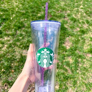 May include: A clear plastic tumbler with a green Starbucks logo and a purple straw. The tumbler has a lid and is held in a hand.