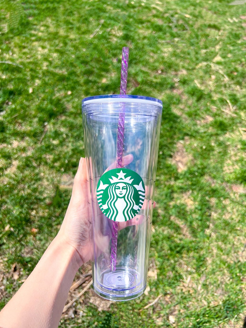May include: A clear plastic tumbler with a green Starbucks logo and a purple straw. The tumbler has a lid and is held in a hand.