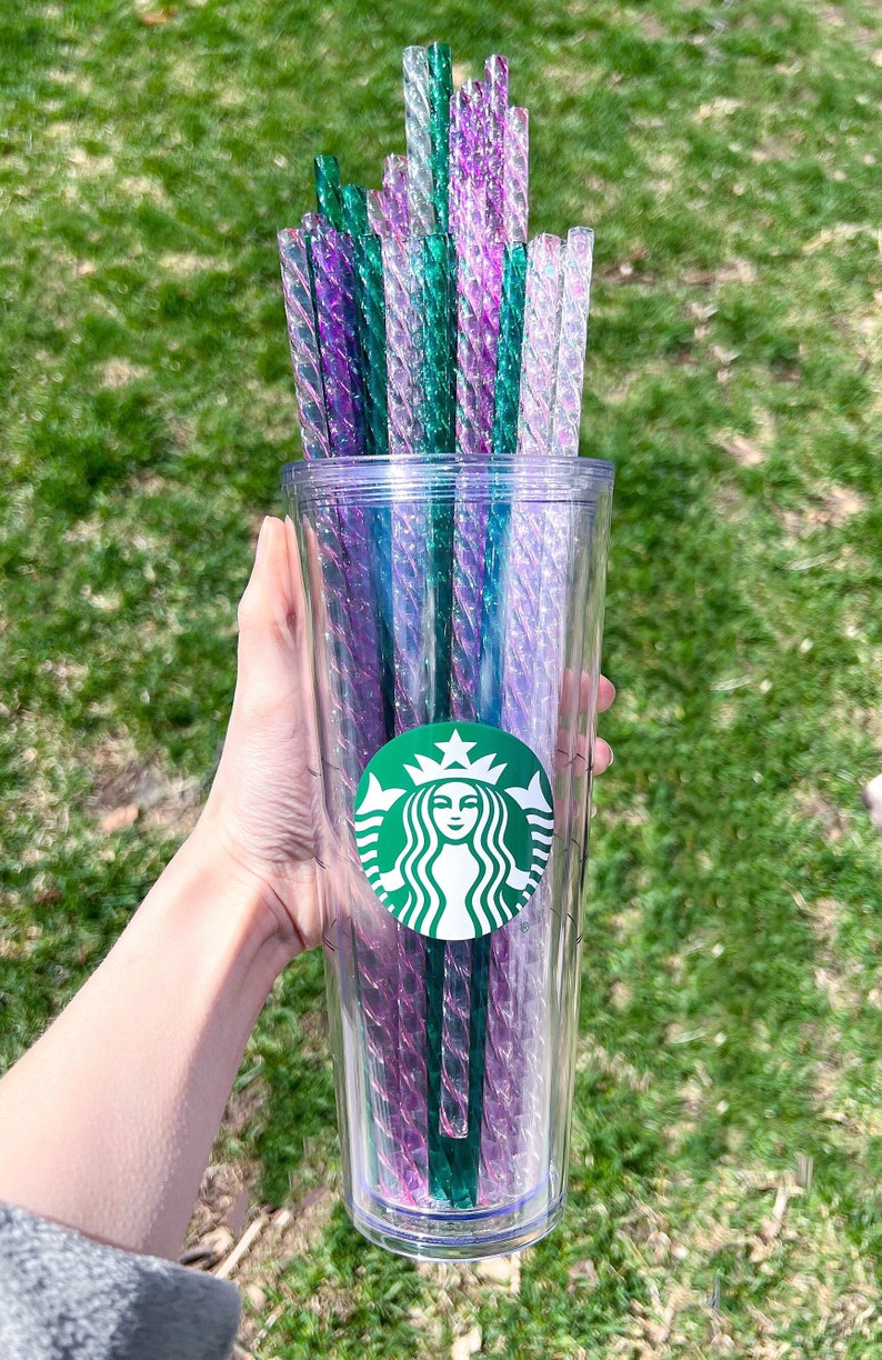 May include: A clear reusable tumbler with a green Starbucks logo and a mermaid design. The tumbler is filled with a bundle of iridescent green and purple straws.