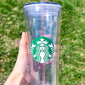 May include: A clear plastic tumbler with a green Starbucks logo and a pink and clear reusable straw. The tumbler has a lid and a measurement scale on the side.