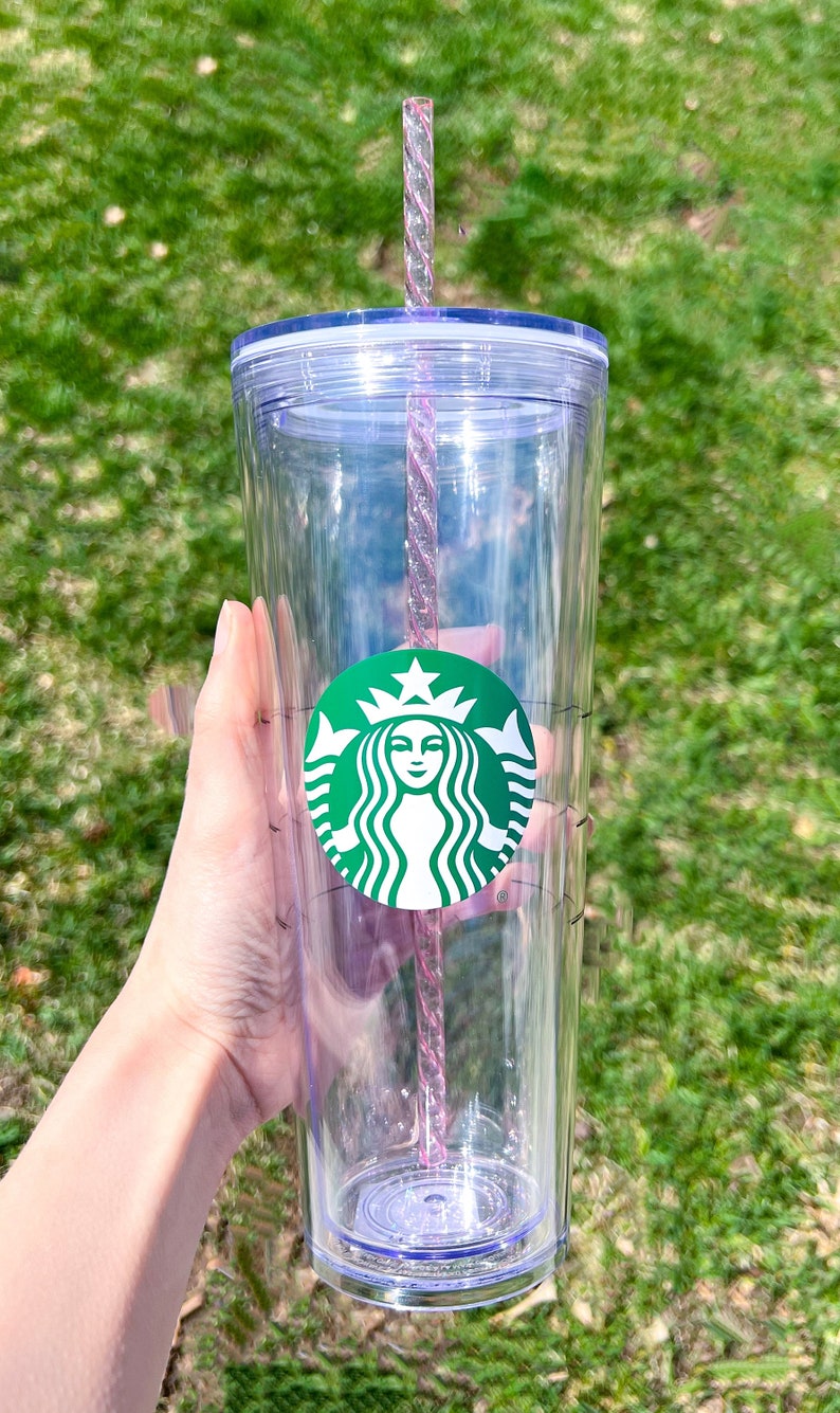 May include: A clear plastic tumbler with a green Starbucks logo and a pink and clear reusable straw. The tumbler has a lid and a measurement scale on the side.