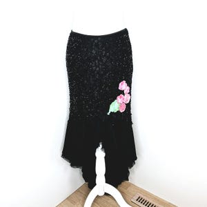 May include: A black skirt featuring a heavily beaded upper section and a smooth, solid black lower hem. The skirt is adorned with a cluster of three-dimensional pink fabric flowers and a single green leaf accent on the right side of the hip area.