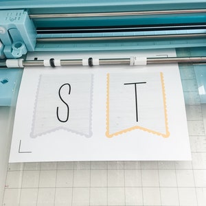 May include: A close-up of a white sheet of paper with the letters "S" and "T" printed on it. The letters are in a banner shape with scalloped edges. The paper is in a cutting machine.