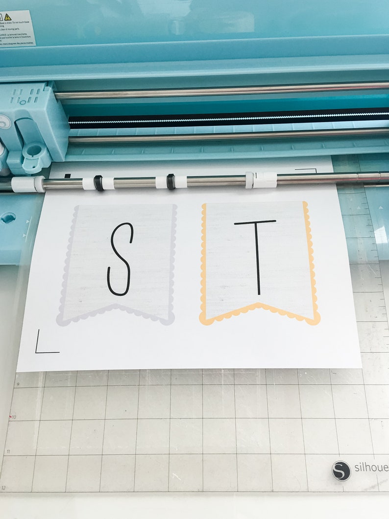 May include: A close-up of a white sheet of paper with the letters "S" and "T" printed on it. The letters are in a banner shape with scalloped edges. The paper is in a cutting machine.