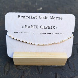 Peut inclure: Bracelet en perles blanches avec des perles dorées, présenté sur une carte blanche avec le texte "Bracelet Code Morse" et "< MAMIE CHERIE >" en noir. Le bracelet est composé de perles blanches et dorées et est enfilé sur un cordon blanc.