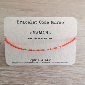 Peut inclure: Un bracelet en perles rouges et dorées avec le mot "MAMAN" en code Morse sur une carte blanche. La carte indique également "Bracelet Code Morse" et "Sophie & Lilli".