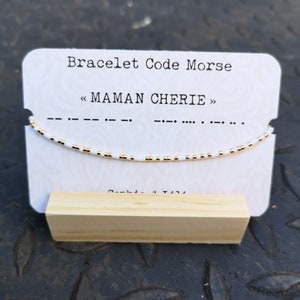 Peut inclure: Bracelet en perles blanches avec des perles dorées, sur une carte blanche avec le texte "Bracelet Code Morse" et "<< MAMAN CHERIE >>" en noir. Le bracelet est sur un support en bois.