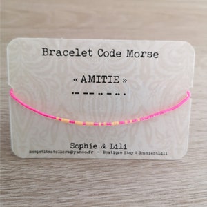 Peut inclure: Bracelet d'amitié rose et orange avec le mot "AMITIE" en code morse sur une carte blanche. La carte indique également "Bracelet Code Morse" et "Sophie & Lili".