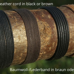 Może przedstawiać: Zbliżenie na cztery sznurki owinięte wok&oacute;ł drewnianego kloca. Sznurki są czarne, brązowe, czarne i brązowe. Tekst "Cotton / leather cord in black or brown" znajduje się w g&oacute;rnej części obrazu. Tekst "Baumwoll-/Lederband in braun oder schwarz" znajduje się w dolnej części obrazu.