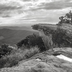 May include: A black and white photograph of a rocky cliff overlooking a mountain range. The cliff is shaped like a bird's beak and extends out over the valley below. The sky is cloudy and the mountains are covered in trees.