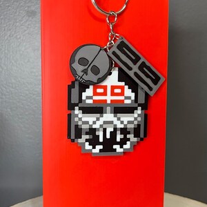 May include: A pixelated skull keyring with a silver ring. The keyring features a grey skull, a grey tag with the number "29", and a pixelated skull design with red accents. The background is a bright red.