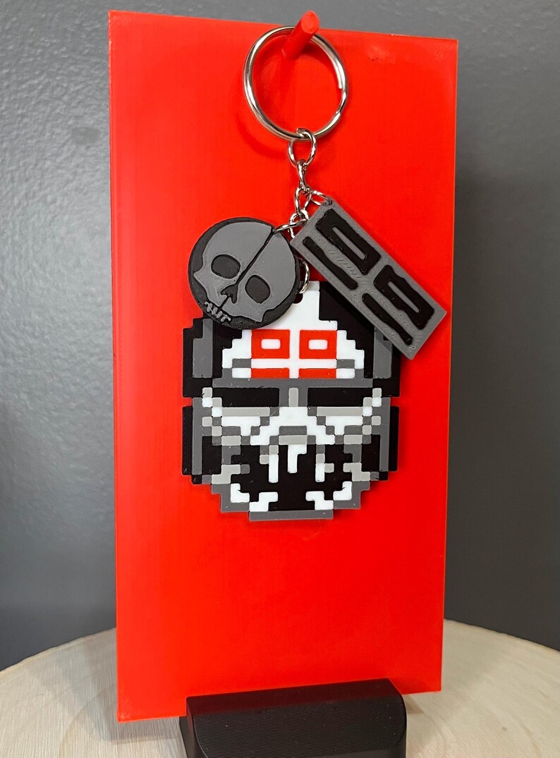 May include: A pixelated skull keyring with a silver ring. The keyring features a grey skull, a grey tag with the number "29", and a pixelated skull design with red accents. The background is a bright red.