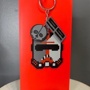 May include: A pixelated keychain with a grey skull and a helmet design with orange and black details. The keychain is attached to a silver keyring, set against a bright red background.