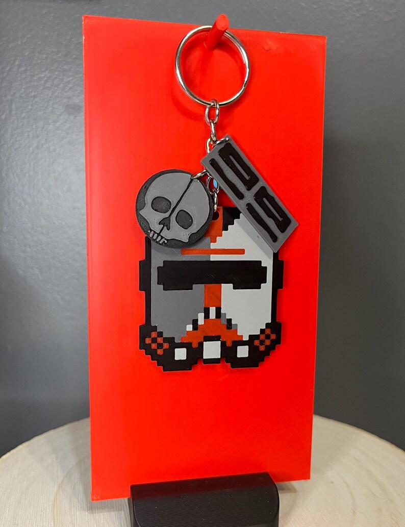 May include: A pixelated keychain with a grey skull and a helmet design with orange and black details. The keychain is attached to a silver keyring, set against a bright red background.