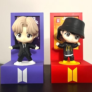 Può includere: Due figurine da collezione, una in abito nero e l'altra in completo nero con logo "M" giallo, sono su espositori viola e rossi. Gli espositori presentano il logo BTS e sono incorniciati.