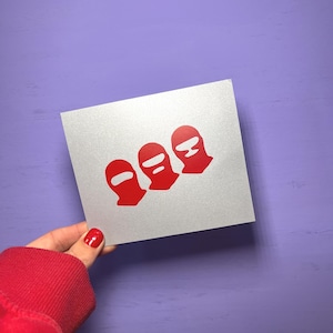 May include: A silver card featuring three red ski mask graphics. The ski mask graphics are arranged in a row. The card is held by a person with red nail polish and a red sleeve.