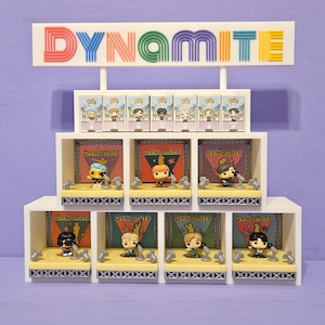 May include: A white display unit featuring the word "DYNAMITE" in vibrant colours. It showcases miniature figures in boxes and on stage-like platforms. Each figure has a unique design, with microphones and stage backdrops.