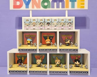 Pode incluir: Uma prateleira branca com a palavra "DYNAMITE" em letras coloridas. A prateleira contém figuras em miniatura em caixas e em plataformas semelhantes a palcos. Cada figura tem uma cor e design diferentes, com microfones e cenários.