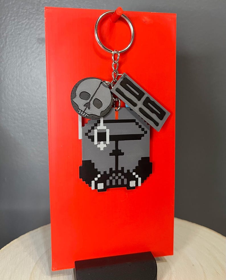 May include: A grey pixelated keychain featuring a skull, a rectangular tag with the number "92", and a pixelated character design. The keychain is attached to a silver keyring and displayed against a bright red background.